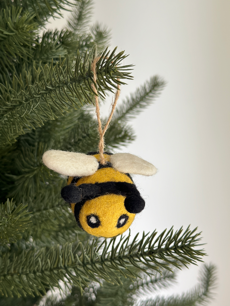 A cheerful felted wool bee ornament hangs from a branch of an evergreen Christmas tree. This handmade ornament features a round yellow and black striped body with small, delicate white wings, suspended by a jute string, adding a touch of handcrafted charm.