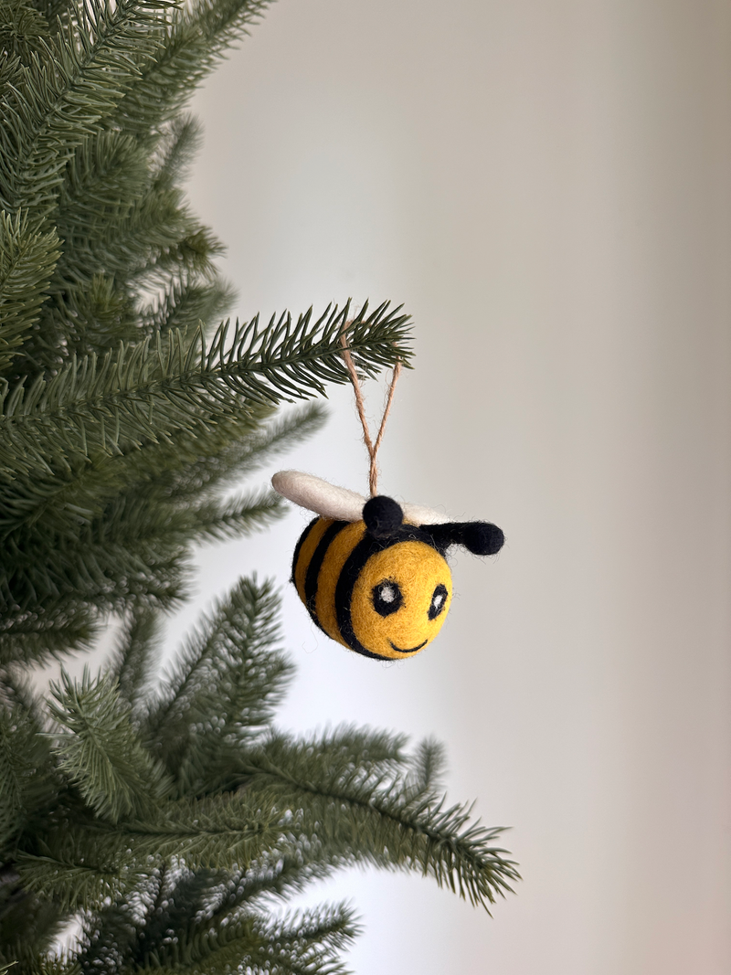 A cheerful wool felt bee ornament hangs from a pine tree branch. This felted ornament features alternating bright yellow and black bands, a smiling face with two black eyes, a stitched smile, small white felt wings, and two black antennae. A jute string is attached to the bee for hanging.