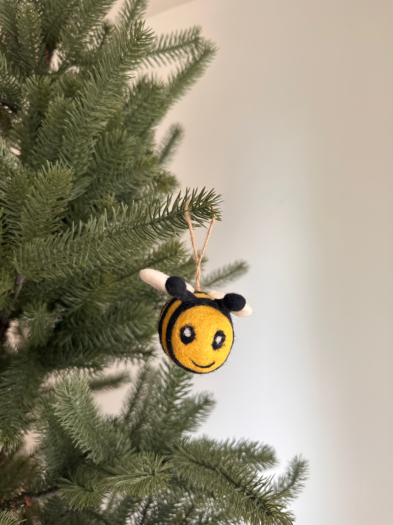 A cheerful felted wool bee ornament hangs gracefully on a lush green Christmas tree, set against a soft off-white wall. This round bee features bright yellow and black stripes, small black antennae, tiny white wings, and a heartwarming stitched smile with oval eyes. A jute string is visible at the top, facilitating its display.