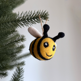 A felt bee ornament hangs on a Christmas tree, creating a joyful, festive mood. This handcrafted bee features a round body with alternating yellow and black stripes. Its face has white and black eyes with a black stitched smile. Two white felt wings extend from its back, and black antennae protrude from its head, with a jute string attached for hanging this unique Christmas ornament.