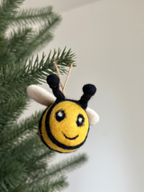 A cheerful felted wool bee Christmas ornament hangs on a green Christmas tree branch. The bee features a round, yellow body with black stripes, small white wings, black antennae, white eyes, and a smiling face. A jute string is attached for hanging.