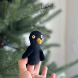 A black raven felt finger puppet is playfully held by a female hand, set against a blurred Christmas tree. The handmade felt raven finger puppet features a black body with a striking yellow beak and bright yellow and white eyes, perfect for whimsical, imaginative play.