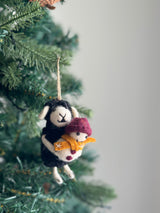 A joyful felt black sheep Christmas ornament hangs on a festive Christmas tree. This unique ornament depicts a black sheep with white accents on its face and ears, gently holding a snowman. The snowman features a maroon hat, orange scarf, and maroon dots, adding vibrant details. A jute string is attached for easy display.
