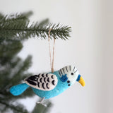 A blue budgerigar felt ornament hangs gracefully from a pine branch, set against a bright, neutral backdrop. This handcrafted felt ornament features detailed needle-felted markings on its head and wings, a bright yellow beak, and a blue tail. A jute string is attached, adding a touch of festive cheer.