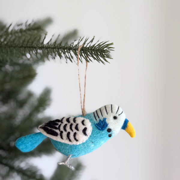 A blue budgerigar felt ornament hangs gracefully from a pine branch, set against a bright, neutral backdrop. This handcrafted felt ornament features detailed needle-felted markings on its head and wings, a bright yellow beak, and a blue tail. A jute string is attached, adding a touch of festive cheer.