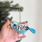 A unique blue Budgerigar felt ornament is displayed against a light, airy backdrop, with a subtle glimpse of a Christmas tree branch. This handcrafted wool felt ornament features a bright blue body, a white head with black markings, and a distinctive yellow beak. It also has unique black and white patterned wings, small gray felt feet, and a jute string for hanging.