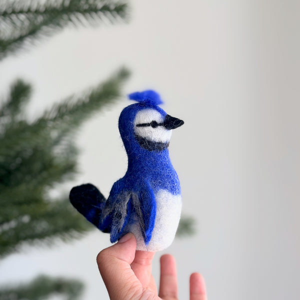 A handcrafted wool felt blue jay finger puppet, made by fair-trade artisans, is gently presented by a female hand against a soft, naturally lit background with a hint of a Christmas tree. This vibrant blue jay felt finger puppet features striking blue feathers on its head, wings, and tail, a distinctive crest, a white belly, and black accents around its beak, highlighting its realistic charm.