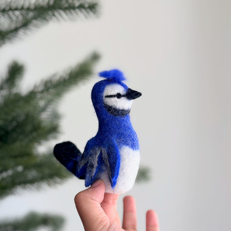 A handcrafted wool felt blue jay finger puppet, made by fair-trade artisans, is gently presented by a female hand against a soft, naturally lit background with a hint of a Christmas tree. This vibrant blue jay felt finger puppet features striking blue feathers on its head, wings, and tail, a distinctive crest, a white belly, and black accents around its beak, highlighting its realistic charm.