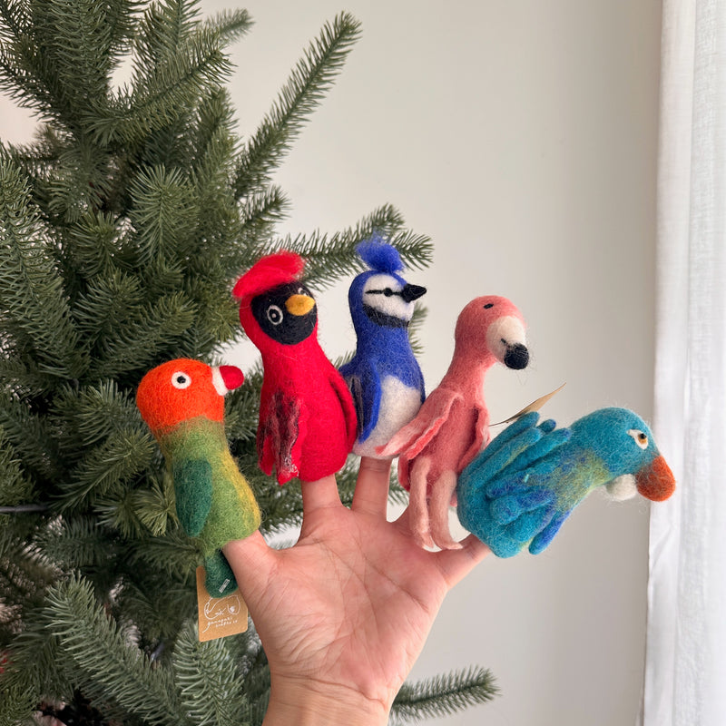 A needle-felted blue jay finger puppet is displayed on a female hand, set against a festive backdrop of a Christmas pine tree and a white curtain. This handcrafted blue jay puppet showcases striking blue feathers on its head, wings, and tail, and a distinctive crest. Its unique felt texture adds to its handmade charm, inviting imaginative play.