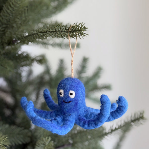 A wool felt blue octopus ornament hangs from a green pine branch against a soft white backdrop, exuding a whimsical and cheerful mood. This vibrant blue felt octopus ornament features eight curly tentacles, small black eyes, and a simple stitched smile. Made from fine wool felt, it offers a soft yet durable texture, ready for hanging by its jute string.