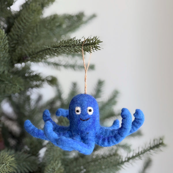 A blue wool felt octopus ornament hangs from a vibrant green pine branch, set against a soft, neutral background. This playful felt octopus features curly tentacles and a cheerful expression with small, round eyes and a stitched smile. A rustic jute string suspends the unique ornament, adding a whimsical touch to the festive scene.