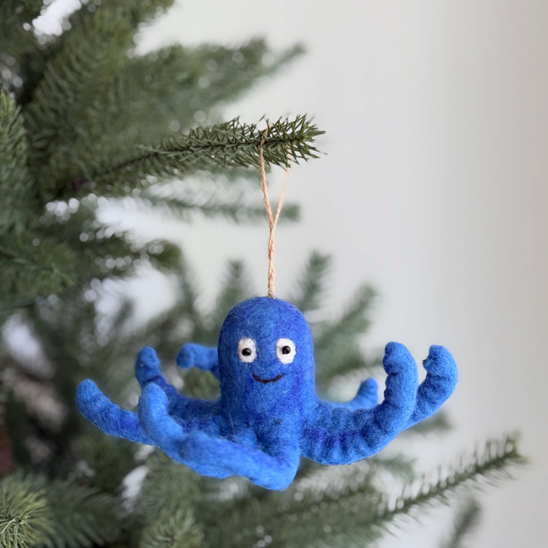 A blue wool felt octopus ornament hangs from a vibrant green pine branch, set against a soft, neutral background. This playful felt octopus features curly tentacles and a cheerful expression with small, round eyes and a stitched smile. A rustic jute string suspends the unique ornament, adding a whimsical touch to the festive scene.
