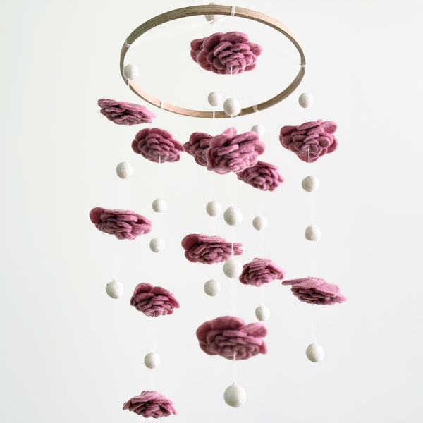A Blush Rose wool felt baby mobile hangs against a plain white background. It features a natural wood hoop from which multiple hand-felted wool flowers in a soft blush rose color alternate with evenly spaced felt pearls. The layered petal design of the flowers creates a delicate and elegant look, enhancing the mobile's calming aesthetic.