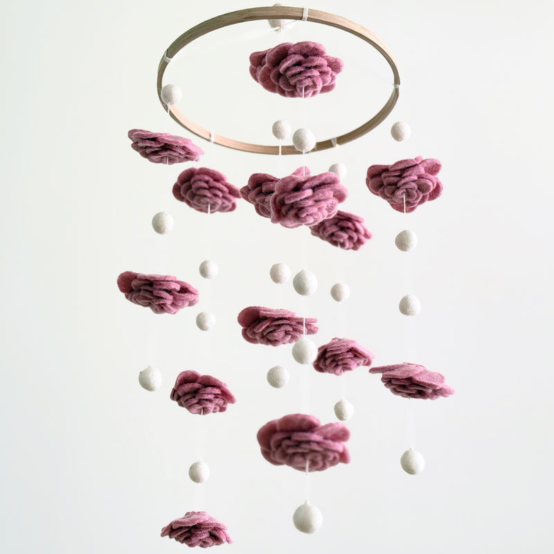A Blush Rose wool felt baby mobile hangs against a plain white background. It features a natural wood hoop from which multiple hand-felted wool flowers in a soft blush rose color alternate with evenly spaced felt pearls. The layered petal design of the flowers creates a delicate and elegant look, enhancing the mobile's calming aesthetic.