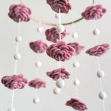 A Blush Rose wool felt baby mobile is presented in a close-up against a muted white background. It features delicate, hand-felted blush-toned wool flowers and felt pearls artfully arranged around a natural wood hoop. The soft wool felt texture and smooth pearls create a gentle, calming aesthetic, highlighting its handcrafted elegance.