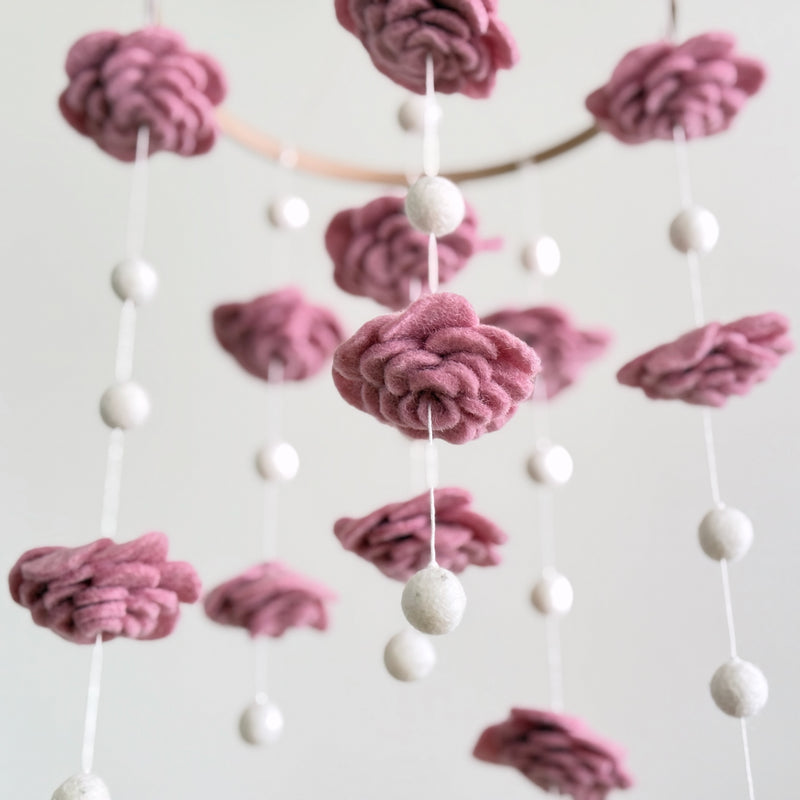 A Blush Rose wool felt baby mobile is presented in a close-up against a muted white background. It features delicate, hand-felted blush-toned wool flowers and felt pearls artfully arranged around a natural wood hoop. The soft wool felt texture and smooth pearls create a gentle, calming aesthetic, highlighting its handcrafted elegance.