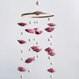 A blush rose wool felt baby mobile is suspended against a soft, neutral backdrop, evoking calm and gentle elegance. Delicate, hand-felted flowers in dreamy pastel pink tones are arranged around a natural wood hoop. White strings and felt pearls dangle from the hoop and between the layers of flowers, creating a visually soothing and romantic accent for a nursery.