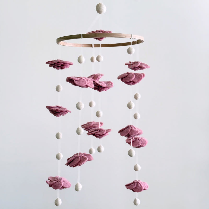 A blush rose wool felt baby mobile is suspended against a soft, neutral backdrop, evoking calm and gentle elegance. Delicate, hand-felted flowers in dreamy pastel pink tones are arranged around a natural wood hoop. White strings and felt pearls dangle from the hoop and between the layers of flowers, creating a visually soothing and romantic accent for a nursery.