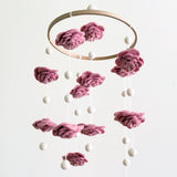 A Blush Rose Flower wool felt baby mobile is gracefully displayed against a clean white background. This enchanting baby mobile features delicate, hand-felted wool flowers in varying shades of pink, artfully resembling blooming roses. Small white felt pearls are suspended between the flowers at different heights from a natural wooden hoop, creating a balanced and dreamy design.