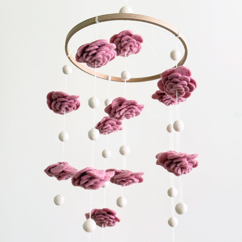 A Blush Rose Flower wool felt baby mobile is gracefully displayed against a clean white background. This enchanting baby mobile features delicate, hand-felted wool flowers in varying shades of pink, artfully resembling blooming roses. Small white felt pearls are suspended between the flowers at different heights from a natural wooden hoop, creating a balanced and dreamy design.