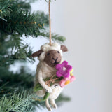 A charming wool felt bouquet-bearing sheep Christmas ornament hangs by a jute string from an evergreen branch, against a softly blurred background. The creamy white sheep features a brown face with textured ears and a stitched nose. It gently holds a vibrant bouquet of pink, magenta, and yellow felt flowers with a green stem.