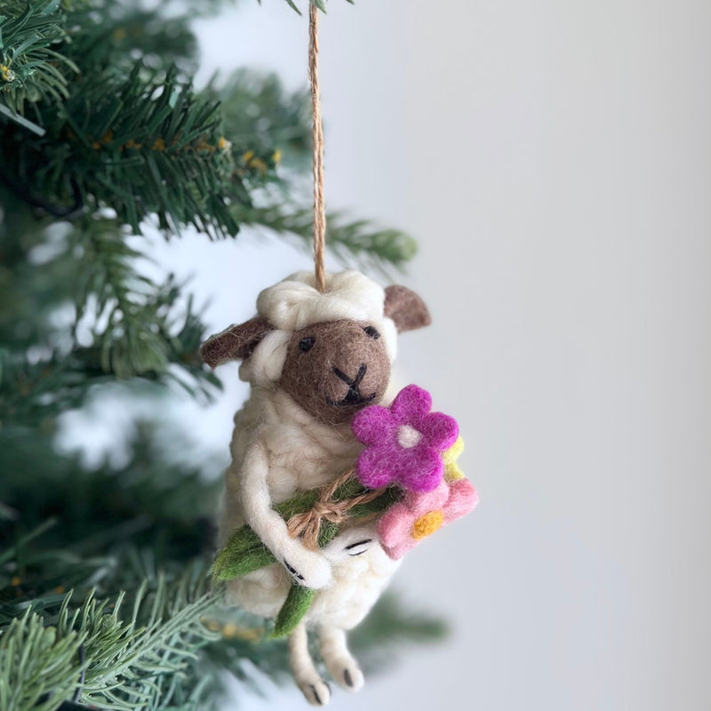 A charming wool felt bouquet-bearing sheep Christmas ornament hangs by a jute string from an evergreen branch, against a softly blurred background. The creamy white sheep features a brown face with textured ears and a stitched nose. It gently holds a vibrant bouquet of pink, magenta, and yellow felt flowers with a green stem.