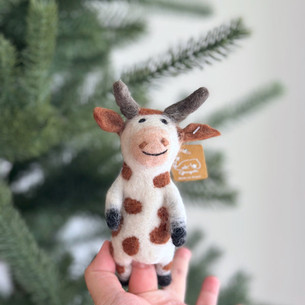 A handmade wool felt Brown Cow finger puppet is held gently in a female hand, set against a softly blurred background of a Christmas pine tree. This charming felt toy features brown spots, ears, and horns. Black hooves and a smiling face add to its playful, handcrafted appeal.