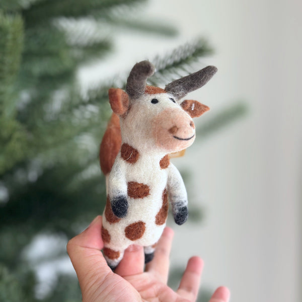 A handmade wool felt brown cow finger puppet is held up by a female hand against a blurred Christmas tree background. This charming puppet is primarily white with distinctive brown spots, ears, and horns. Its gentle expression is created with needle-felted details and a black stitched smile.