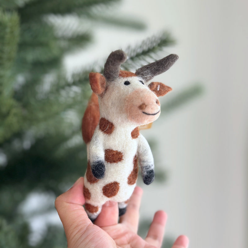 A handmade wool felt brown cow finger puppet is held up by a female hand against a blurred Christmas tree background. This charming puppet is primarily white with distinctive brown spots, ears, and horns. Its gentle expression is created with needle-felted details and a black stitched smile.