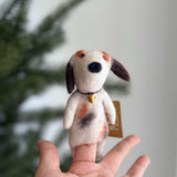 A charming wool felt Calico Dog finger puppet is held aloft on a female hand, set against a softly blurred Christmas pine tree backdrop. This adorable handcrafted felt dog features a white body with brown and black calico patches, floppy brown ears, a black nose, and expressive needle-felted facial details. A tiny gold-colored bell adorns its neck, highlighting its unique charm.