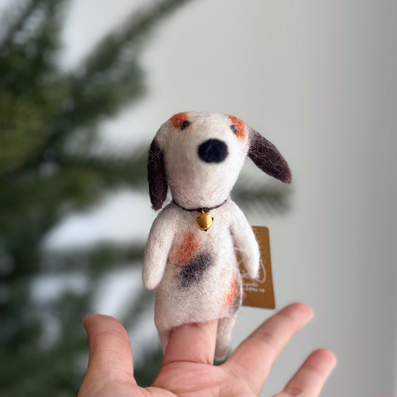 A charming wool felt Calico Dog finger puppet is held aloft on a female hand, set against a softly blurred Christmas pine tree backdrop. This adorable handcrafted felt dog features a white body with brown and black calico patches, floppy brown ears, a black nose, and expressive needle-felted facial details. A tiny gold-colored bell adorns its neck, highlighting its unique charm.