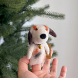 A handcrafted wool felt calico dog finger puppet is held gently in a hand against a blurred Christmas tree background. This charming unique felt finger puppet, known for its fair-trade craftsmanship, features a patchwork of white, orange, and brown spots, a black nose, and floppy brown ears. A tiny golden bell hangs around its neck, adding to its playful and festive appeal.