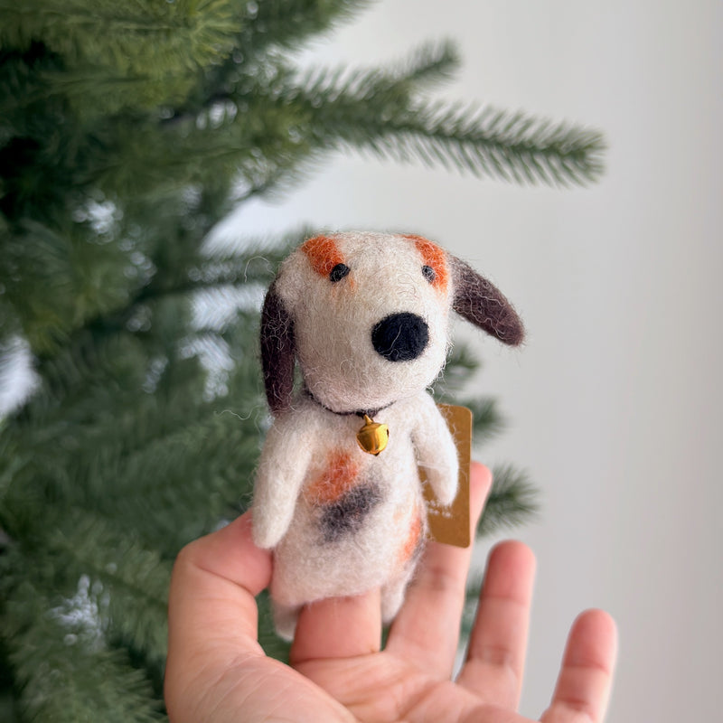 A handcrafted wool felt calico dog finger puppet is held gently in a hand against a blurred Christmas tree background. This charming unique felt finger puppet, known for its fair-trade craftsmanship, features a patchwork of white, orange, and brown spots, a black nose, and floppy brown ears. A tiny golden bell hangs around its neck, adding to its playful and festive appeal.