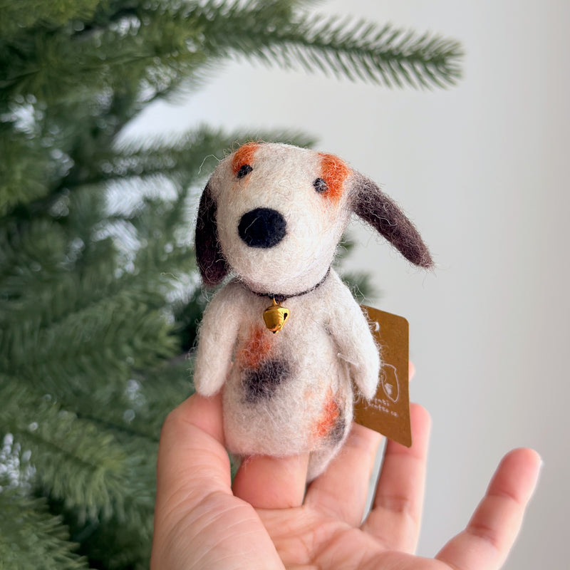 A handmade wool felt Calico Dog Finger Puppet is held in a female hand, set against a blurred Christmas tree backdrop. The unique felted finger puppet features a patchwork coat with white, brown, and orange markings. A small golden bell adorns its neck, adding to its charming appeal.