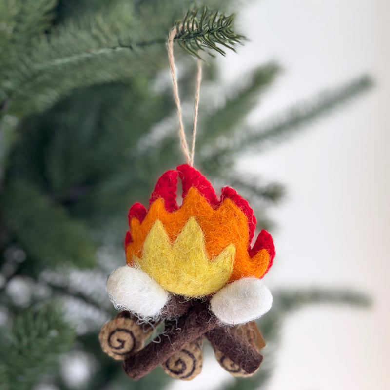 A cozy felt campfire marshmallow ornament hangs from a green Christmas tree branch. This unique Christmas ornament features layered red, orange, and yellow felt flames with three white felted marshmallows toasted over brown felt logs, detailed with spiral patterns, evoking a festive atmosphere.