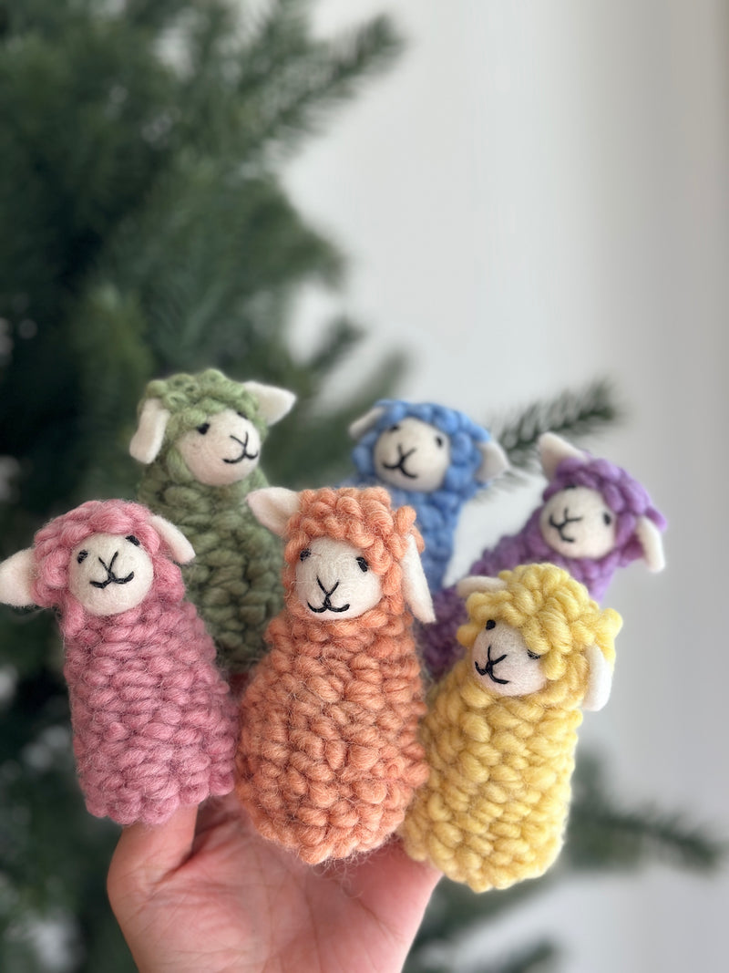 Six pastel felt sheep finger puppets, known as 'Candy Sheep', are held in a hand against a soft, natural backdrop featuring a Christmas tree. Each handmade felt sheep finger puppet, colored pink, green, orange, blue, purple, or yellow, boasts a soft, fluffy 3D design. They feature small white felt ears and a simple, endearing embroidered expression, perfect for imaginative play.