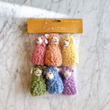 A set of six colorful Candy Sheep felt finger puppets is displayed on a marbled white surface, packaged in a clear plastic bag with a tan cardboard header. These handmade felt finger puppets feature vibrant pink, orange, yellow, green, blue, and purple sheep. Each soft wool felt sheep finger puppet has a textured, wool-like appearance, a white tail, and a simple stitched smiling face, ideal for creative play.