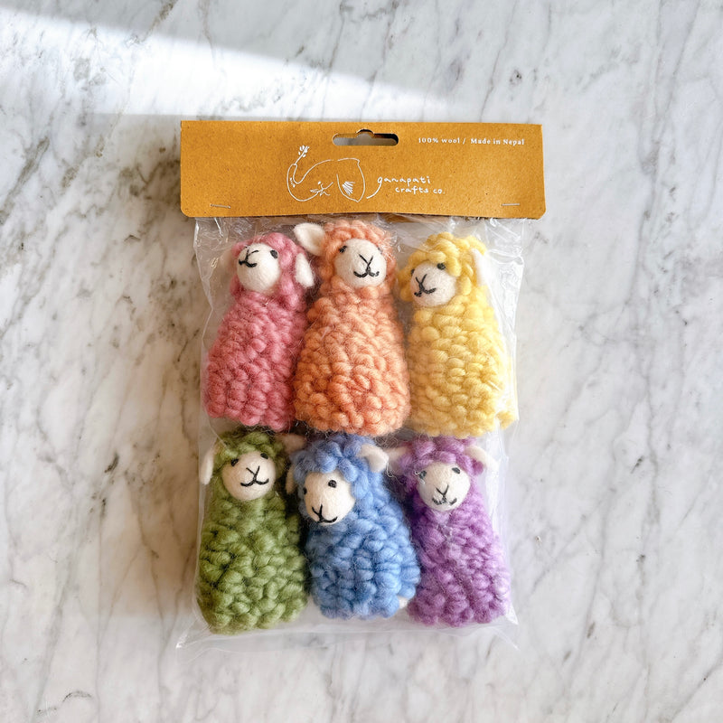 A set of six colorful Candy Sheep felt finger puppets is displayed on a marbled white surface, packaged in a clear plastic bag with a tan cardboard header. These handmade felt finger puppets feature vibrant pink, orange, yellow, green, blue, and purple sheep. Each soft wool felt sheep finger puppet has a textured, wool-like appearance, a white tail, and a simple stitched smiling face, ideal for creative play.