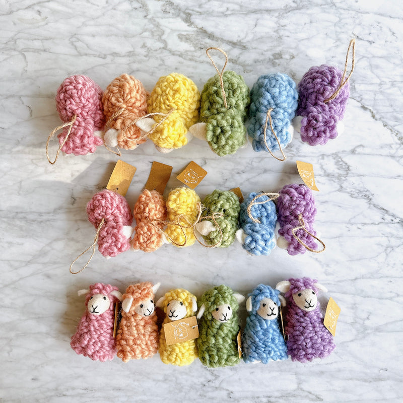 A set of six handcrafted felt sheep finger puppets is displayed in three rows on a marbled surface. Each unique wool felt sheep finger puppet, showcasing its craftsmanship, features a fluffy body, a small smiling face with white ears, and a loop with a tag. They come in soft pastel pink, orange, yellow, green, blue, and purple, creating a visually appealing spectrum.