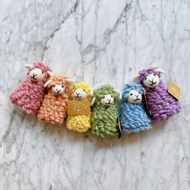 Six handmade wool felt Candy Sheep finger puppets, a charming set of 6, are neatly aligned on a light marble backdrop. Each fluffy wool felt sheep comes in a distinct color: pink, peach, yellow, green, blue, or purple. They feature small white felt ears and simple stitched faces with black mouths.