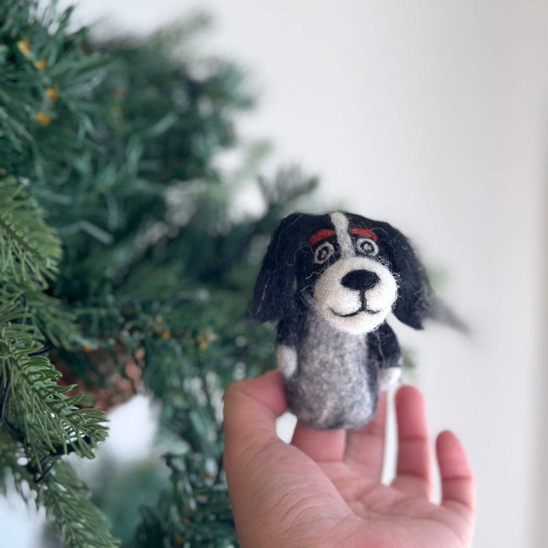 A black Cavalier King Charles Spaniel felt finger puppet is held gently in a hand, set against the cozy, blurred background of a Christmas tree. This handmade felt finger puppet is primarily black with a white face and distinctive red eyebrows. It features fluffy, long, floppy black ears and has a soft texture, perfect for festive finger play.