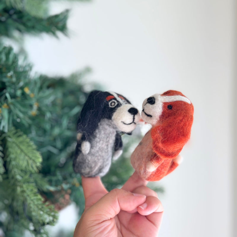 A black Cavalier King Charles Spaniel felt finger puppet is displayed on a hand, set against a blurred Christmas pine tree background, evoking a festive mood. This handmade felt finger puppet features long black ears and a white and gray body, showcasing its soft texture and unique design.