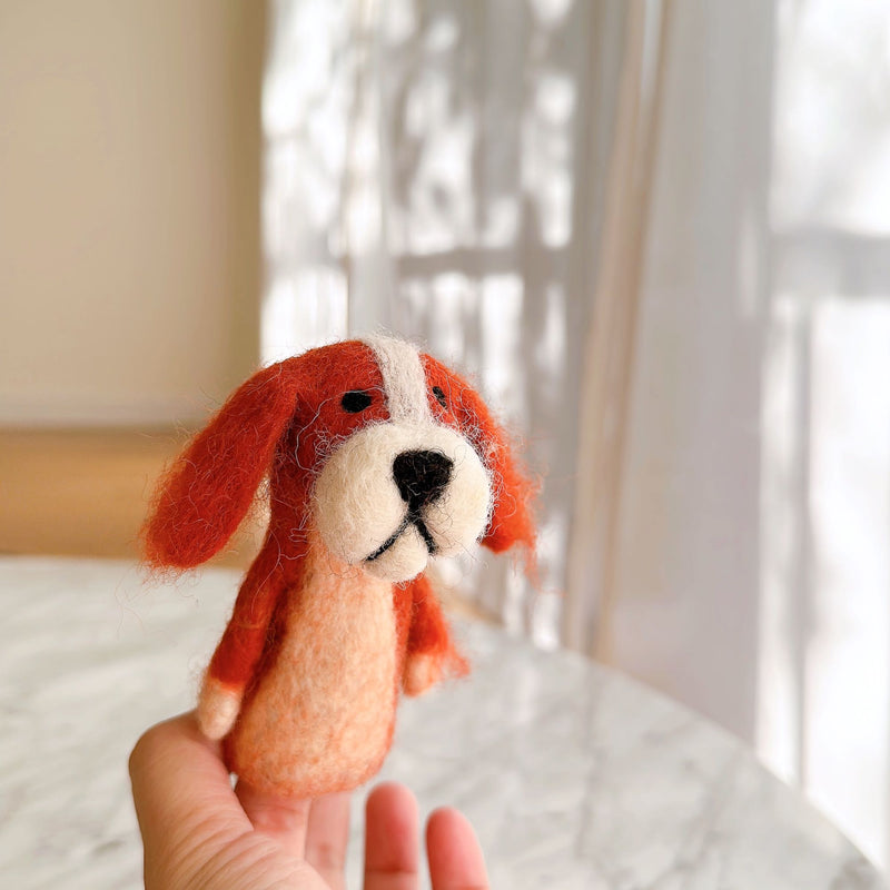 An orange and white Cavalier King Charles Spaniel felt finger puppet is held in a female hand, basking in gentle sunlight. This handmade felt puppet features long fluffy ears, a rounded face with a white stripe, and a soft, textured body, capturing the charming essence of the breed. Soft natural light filters through sheer curtains in the background, enhancing its cozy appeal.