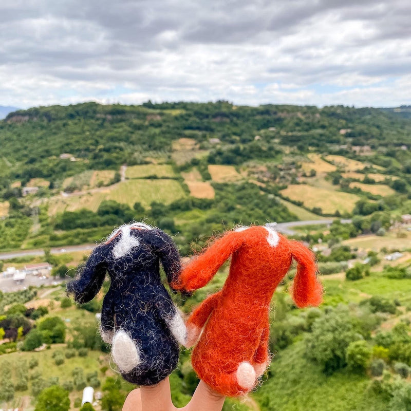 An orange handmade felt Cavalier King Charles Spaniel finger puppet is held in a female hand, against rolling hills and a cloudy sky. This charming, handcrafted felt finger puppet showcases a vibrant orange color, fluffy texture, detailed long ears, and soft facial features. Its unique artistry reflects its fair-trade origin.