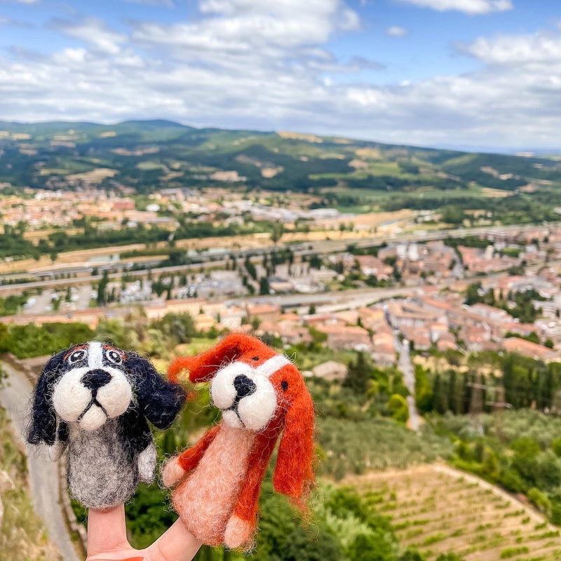 A handmade orange Cavalier King Charles Spaniel wool felt finger puppet is held up by a hand, set against a bright, airy backdrop of rolling hills and a sprawling town. This unique needle felted finger puppet showcases a white marking on its head and floppy ears, featuring a soft, textured appearance characteristic of its quality craftsmanship.