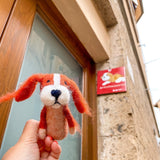 An orange Cavalier King Charles Spaniel wool felt finger puppet is held up close by a female hand. This handcrafted dog puppet features long, fluffy orange ears, a distinct white patch on its head, and a dark blue nose. It is presented outdoors with a building, a wooden door, and a sign depicting a similar dog breed visible in the background, creating a playful scene.