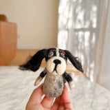 A needle felted Cavalier King Charles Spaniel finger puppet is held by a female hand against a bright, airy background. This handcrafted felt toy features long, fluffy orange ears and a soft, textured orange and white body. Its white face has a small black nose.