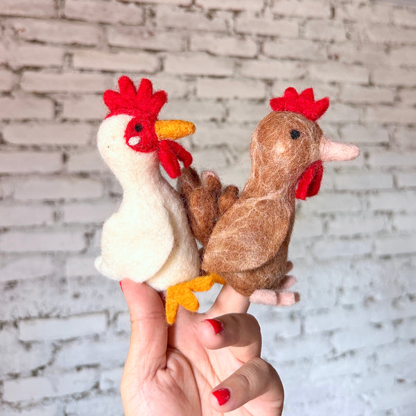Two meticulously handcrafted wool felt chicken finger puppets are held on a female hand against a white brick wall, creating a playful scene. One is a white chicken with a red crest, red wattle, and yellow beak and feet. The second is a brown chicken finger puppet featuring a red wattle and light pink feet, showcasing unique, vibrant details.