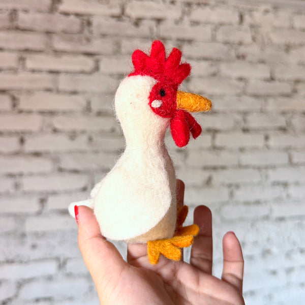 A handmade wool felt chicken finger puppet is held upright in a female hand with red nail polish, set against a blurred white brick background. This uniquely designed chicken features a white body, tiny black eyes, orange beak and feet, and a bright red comb and wattle, showcasing meticulous craftsmanship.