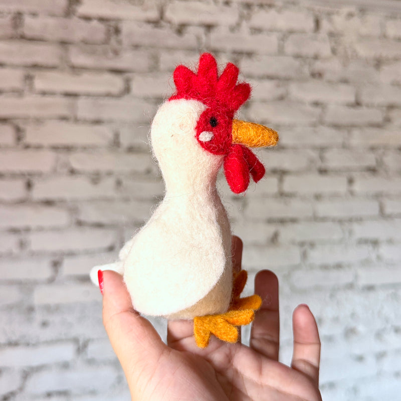 A handmade wool felt chicken finger puppet is held upright in a female hand with red nail polish, set against a blurred white brick background. This uniquely designed chicken features a white body, tiny black eyes, orange beak and feet, and a bright red comb and wattle, showcasing meticulous craftsmanship.